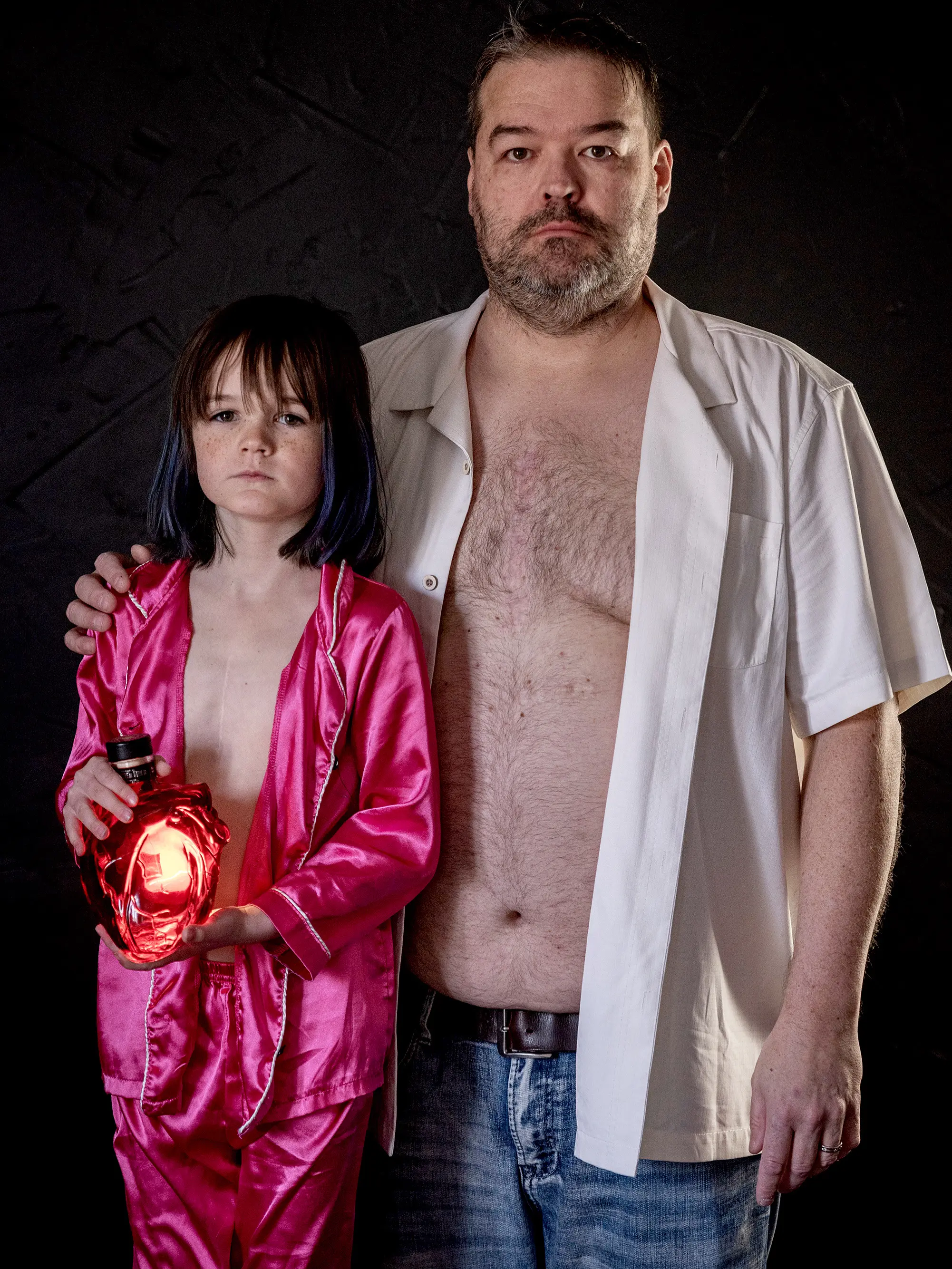 Brooklyn Mitchell, age 8, and her father Joseph Mitchell stand shoulder to shoulder with shirts unbuttoned, revealing nearly identical vertical sternotomy scars, holding an illuminated glass heart between them.